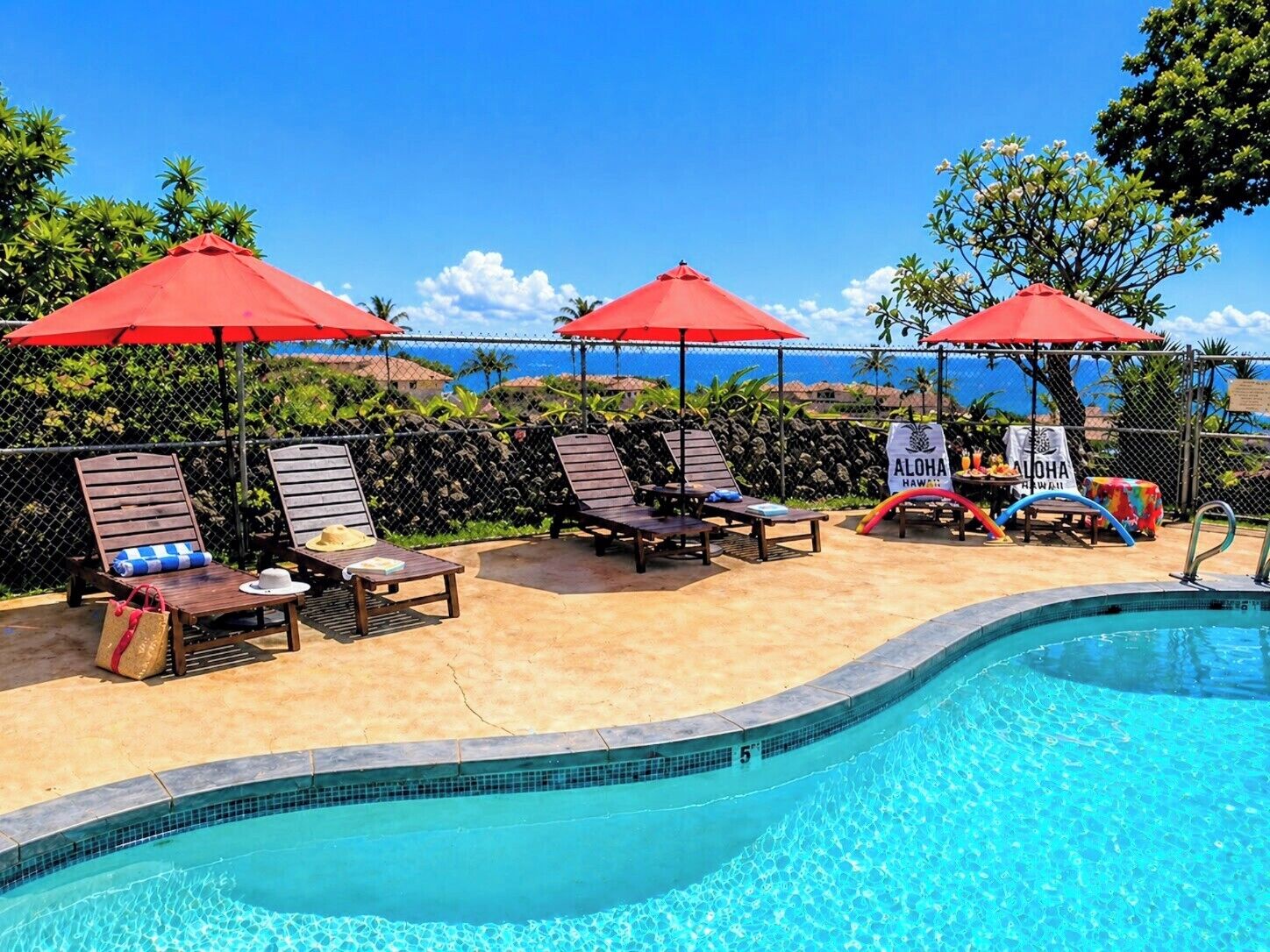 A poolside area with lounge chairs, red umbrellas, and a sign reading 'ALOHA HAWAII'.