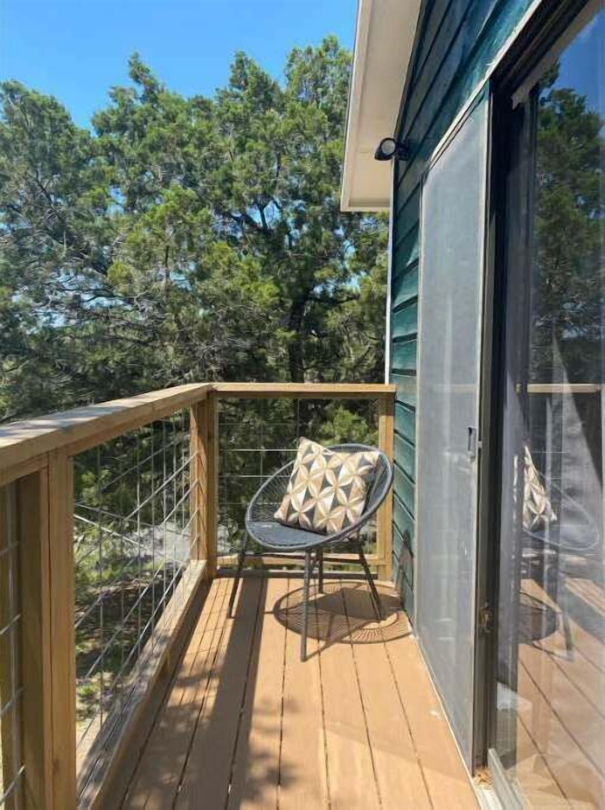 A wooden deck with a glass door leading to a balcony, a chair with a patterned cushion, and trees in the background.