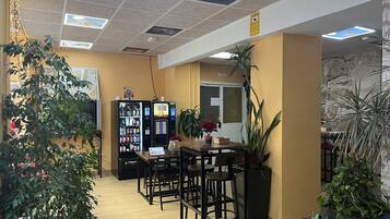 Un restaurante con mesa y sillas de madera, máquinas expendedoras y plantas en macetas.