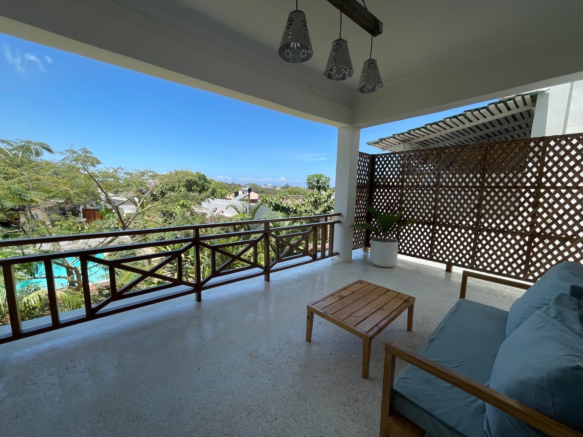 A balcony with a wooden table and a sofa, a view of a pool, and a wooden lattice partition.