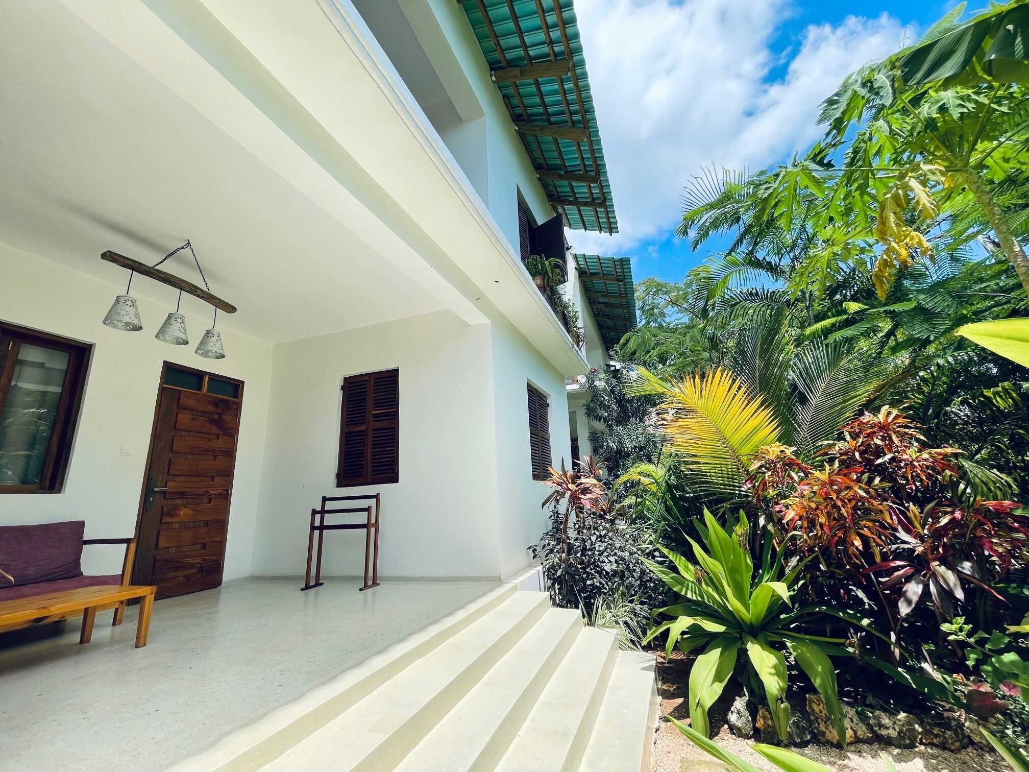 A modern house with a covered porch, a wooden bench, and a variety of tropical plants.