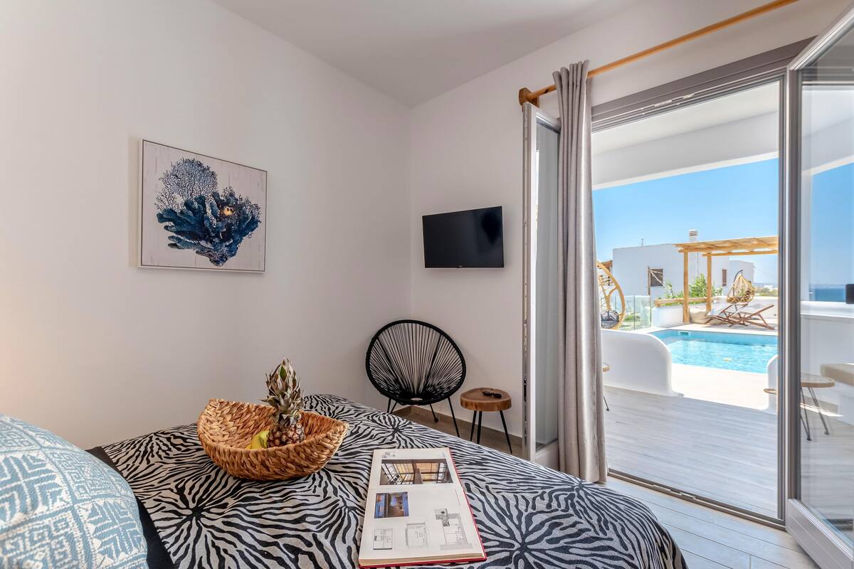 A bedroom with a bed, a wall-mounted TV, a window with a view of a pool and outdoor seating, and a framed artwork on the wall.