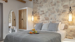 Un dormitorio con pared de piedra, vigas de madera, cama con almohadas estampadas, mesita de noche con una bandeja de frutas y una lámpara colgante.