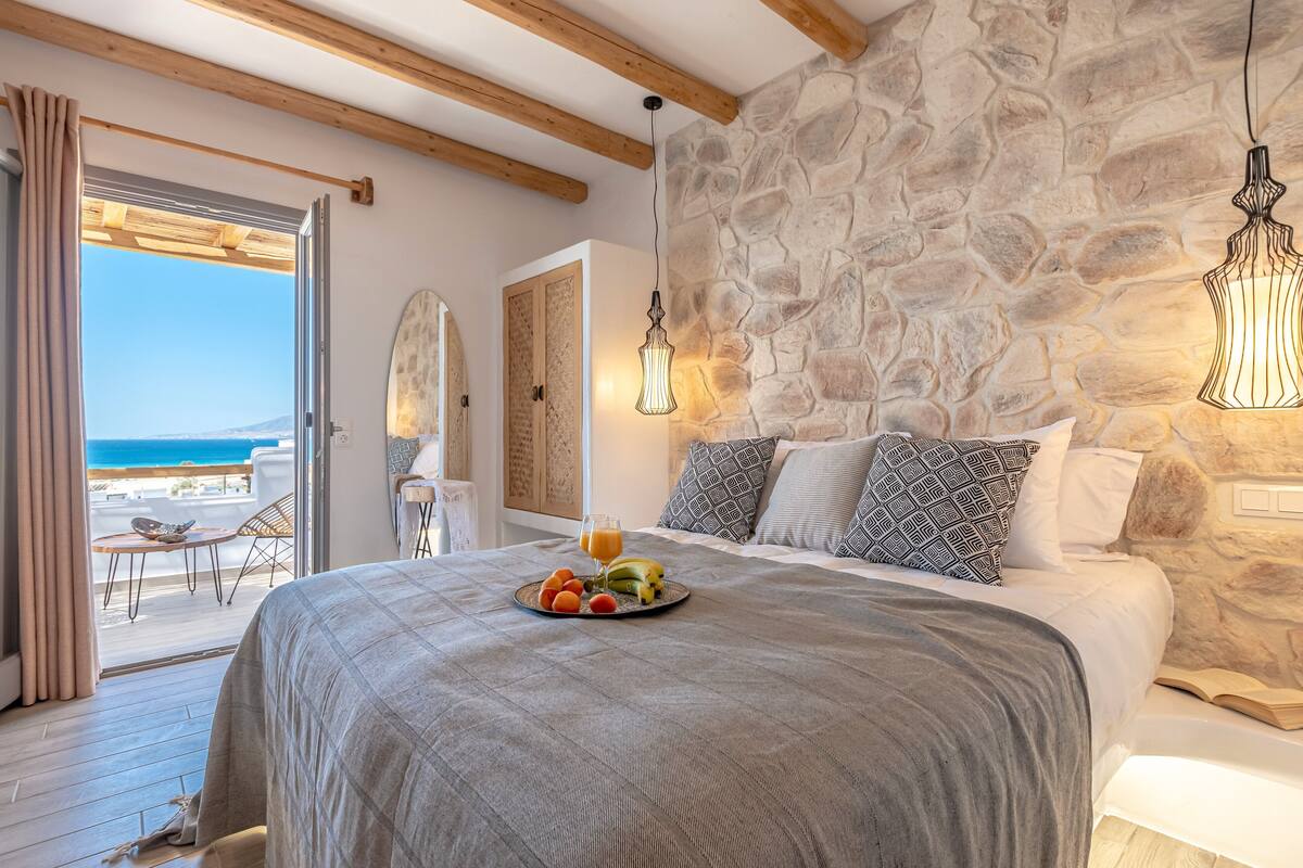 A bedroom with a stone wall, a bed with a fruit plate, and a view of the sea.