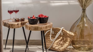 Un balcón con vista a la playa, una mesa de madera con dos copas de vino tinto, un bolso tejido y un jarrón con plumas de plumas.