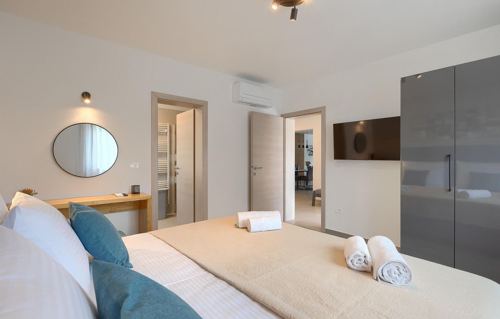 Une chambre d’hôtel moderne, équipée d’un lit, d’une table de chevet, d’un miroir, d’une armoire et d’une salle de bain visible à travers une porte ouverte.