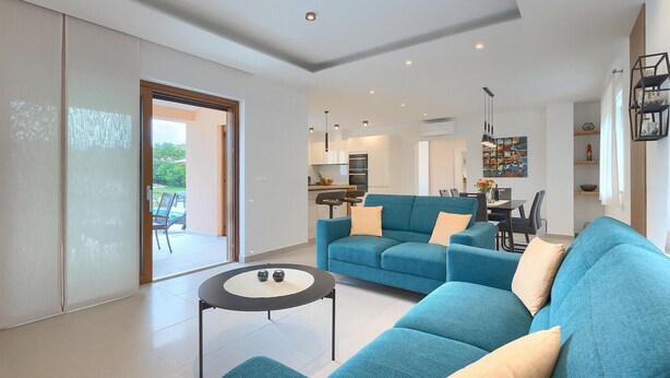 Ein modernes Wohnzimmer mit einem petrolfarbenen Sofa, einem kleinen runden Tisch und einer Küche im Hintergrund.