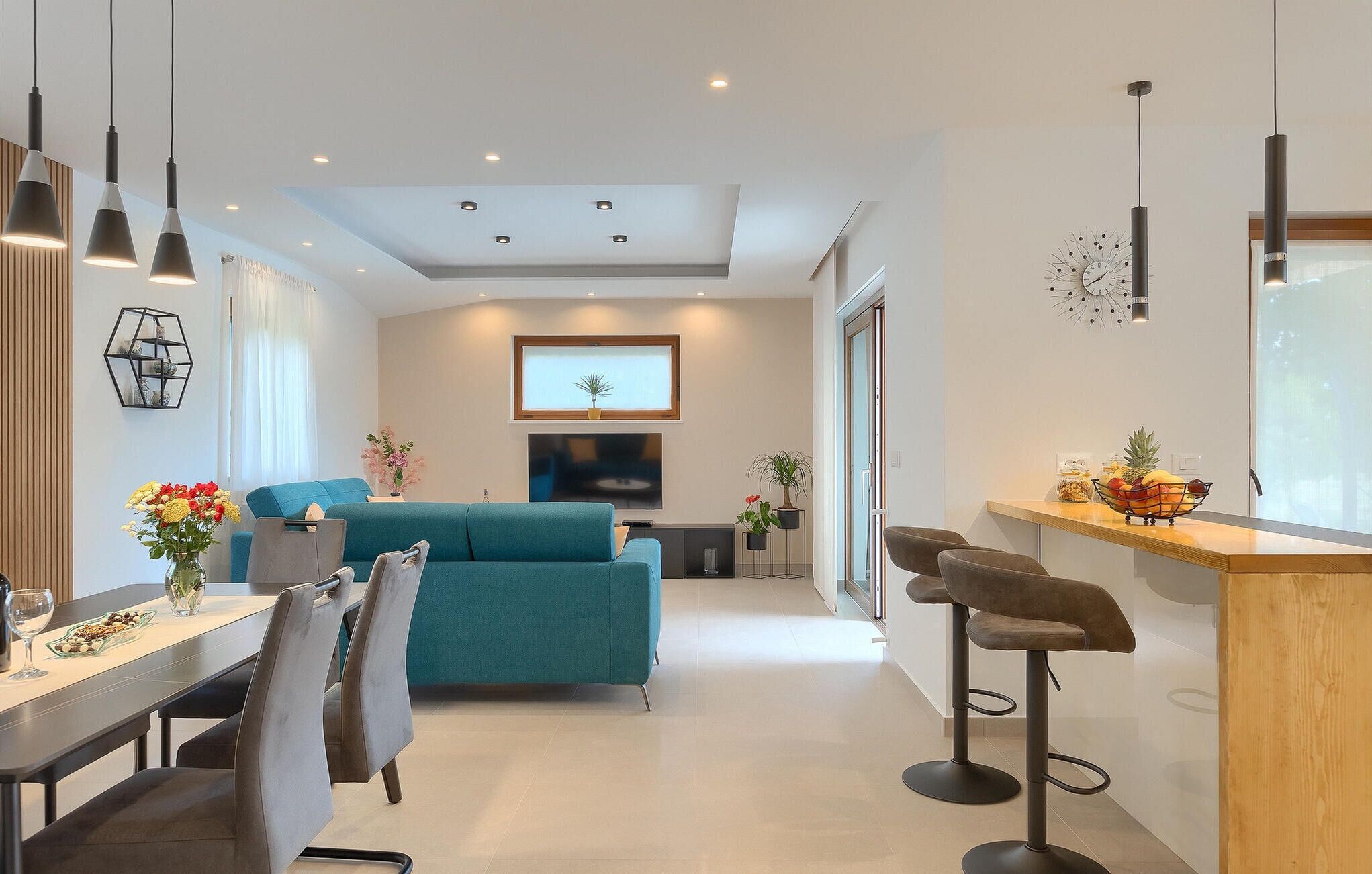 A modern living room with a teal sofa, a dining area with a wooden table and chairs, and a kitchen area with a bar counter.