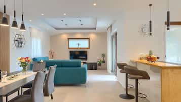 A modern living room with a teal sofa, a dining area with a wooden table and chairs, and a kitchen area with a bar counter.