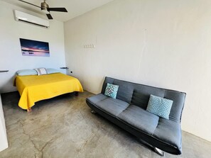 A room with a bed, a sofa, an air conditioner, a ceiling fan, and a wall-mounted light switch.