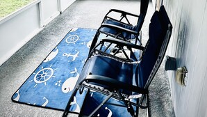 Une terrasse couverte avec un tapis bleu orné de motifs marins, deux chaises noires et un ventilateur de plafond.
