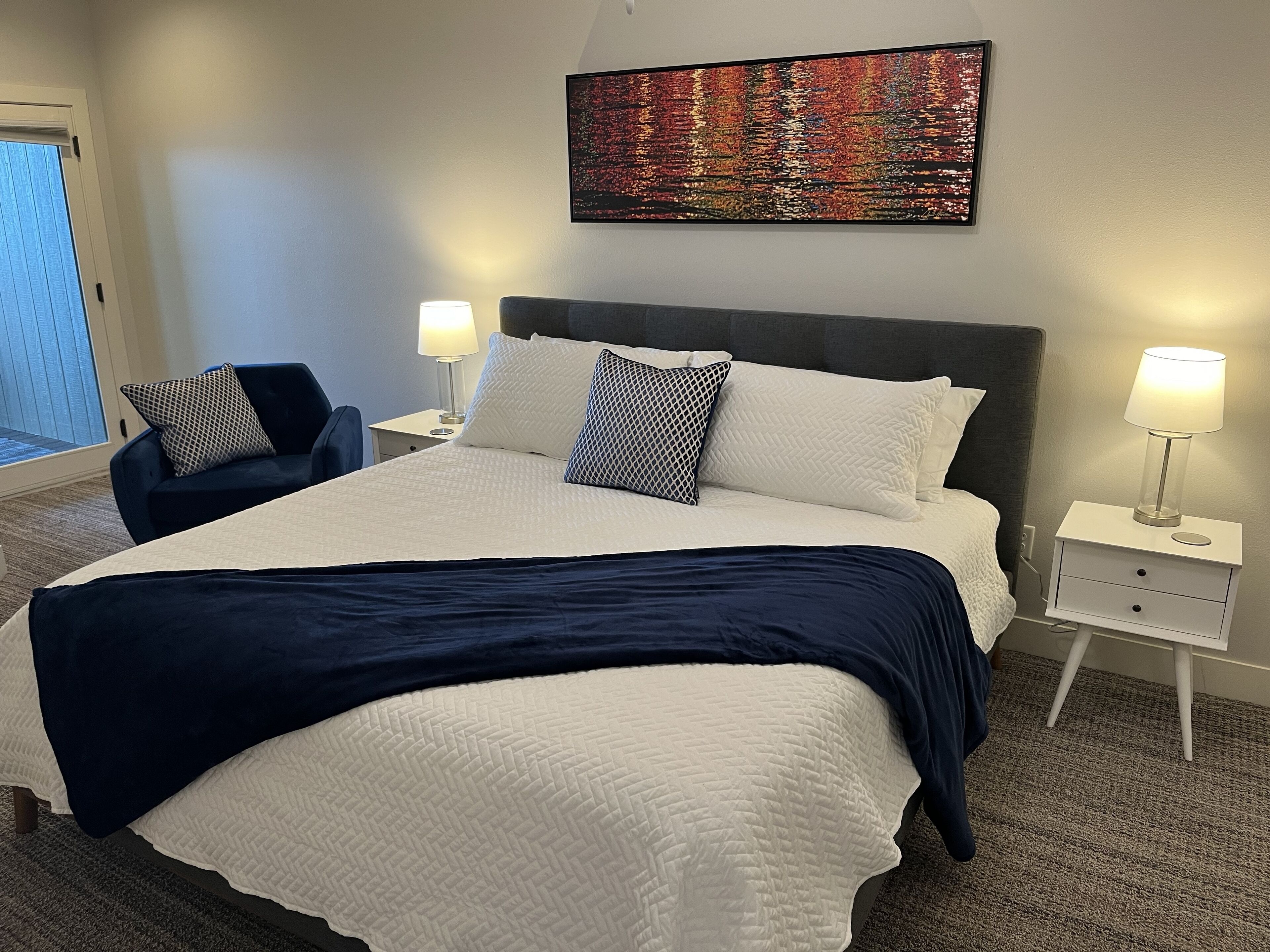 Un lit bien fait, recouvert d’une couvre-lit blanche et bleu marine, avec deux oreillers assortis, et une table de chevet sur laquelle se trouve une lampe. Une chaise est placée au pied du lit, et un tableau abstrait aux couleurs vives est accroché au mur au-dessus.