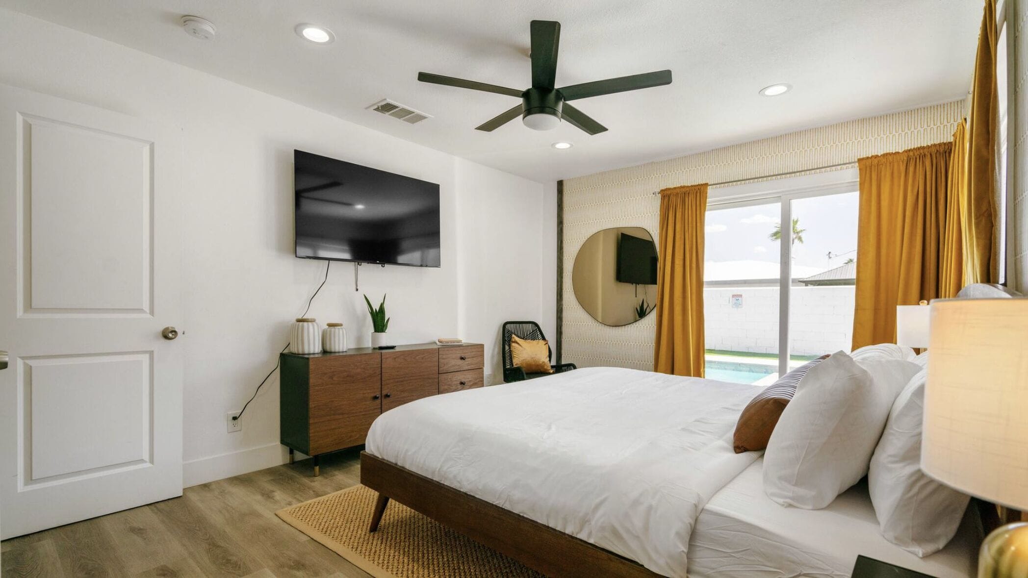 A bedroom with a bed, a TV mounted on the wall, a ceiling fan, a dresser, a mirror, and a window with curtains.