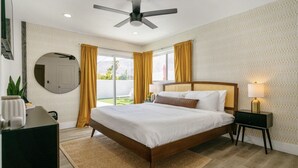 Een slaapkamer met een bed, een plafondventilator, een nachtkastje, een lamp, een spiegel en uitzicht op de omgeving.