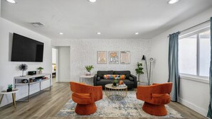 Een moderne woonkamer met een grijze bank, oranje fauteuils, een salontafel en een televisie aan de muur.