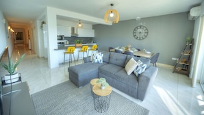 Una sala moderna con un sofá seccional gris, un comedor con sillas amarillas y una cocina al fondo.