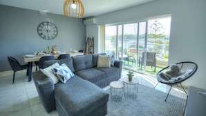 Una sala de estar moderna con un sofá gris, una mesa de comedor para cuatro personas, un ventanal con vista a un puente y una lámpara colgante.
