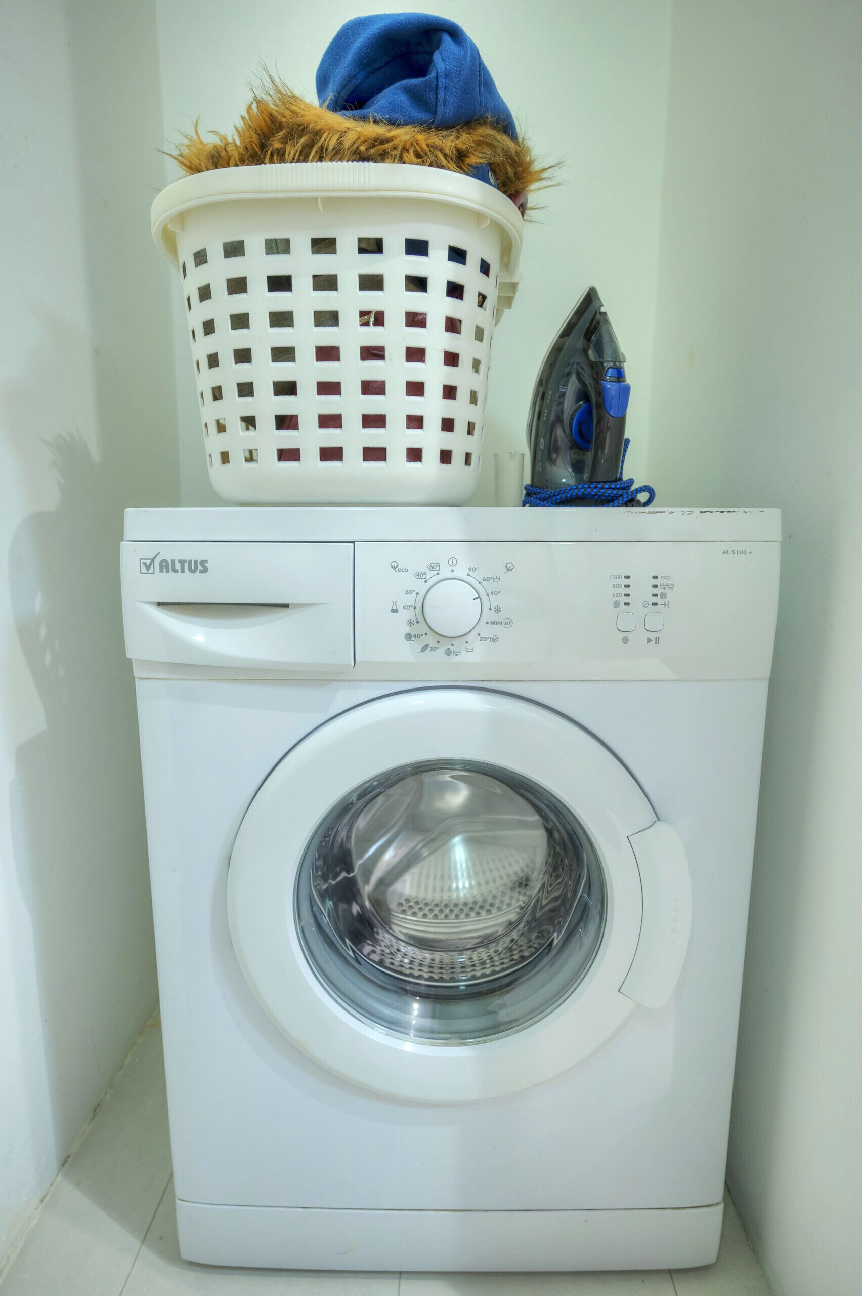 Een voorlader wasmachine met een bedieningspaneel, een mand met een handdoek, en een strijkijzer erboven.