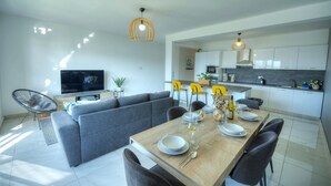 Una sala de estar moderna con un sofá gris, una mesa de comedor de madera preparada para cuatro comensales, y una cocina al fondo.