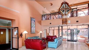 Eine großzügige Lobby mit hoher Decke, einem zentral platzierten roten und blauen Sofa, einer hölzernen Kronleuchter und Blick durch große Glasüren.
