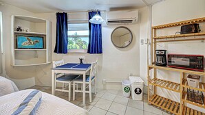 Une pièce de taille modeste comprenant un lit, une petite table avec des chaises, une fenêtre avec des rideaux, un climatiseur fixé au mur, une étagère avec un tableau, un miroir, une poubelle et un four à micro-ondes posé sur une étagère en bois.
