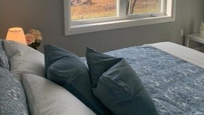 A bedroom with a large window, a bed with patterned bedding, and a lamp on the nightstand.