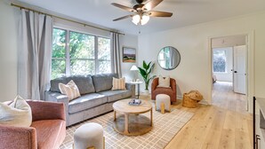 Ein Wohnzimmer mit einer Couch, Sesseln, einem Couchtisch, einem Deckenventilator, einem großen Fenster und einer Tür zu einem anderen Raum.