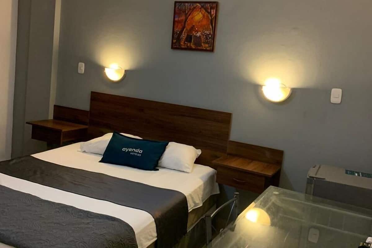 Una habitación de hotel con cama, mesitas de noche, una mesa de vidrio, ventilador de techo y luces fijadas en la pared.