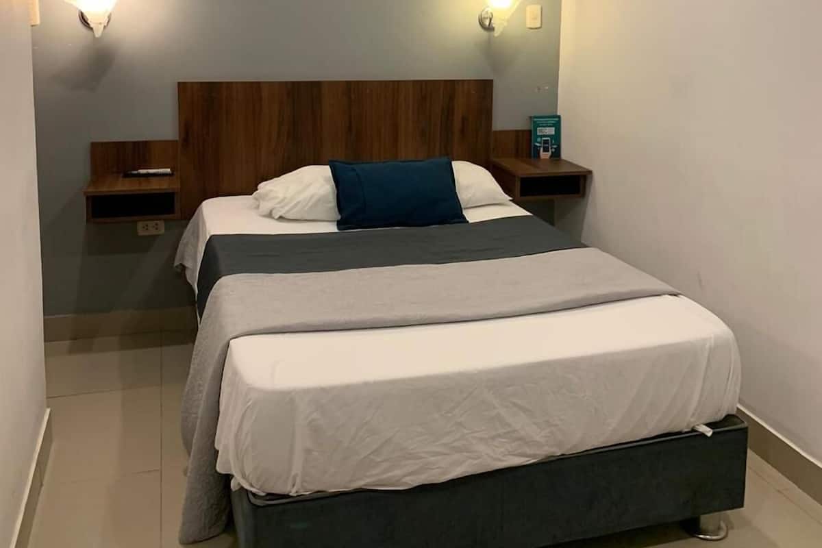 Un dormitorio con cama doble, ventilador de techo, dos mesitas de noche con lámparas, una mesita auxiliar y un cuadro colgado en la pared.