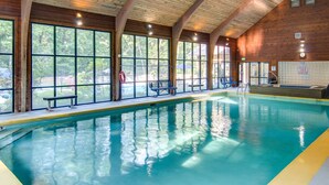 Une grande piscine intérieure à l’eau d’un bleu limpide, avec des poutres en bois au plafond et de grandes fenêtres offrant une vue sur l’extérieur.