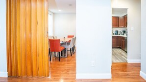 Ein Zimmer mit Holzboden, einem Esstisch mit Stühlen und einer durch einen offenen Durchgang erreichbaren Küchenzeile.