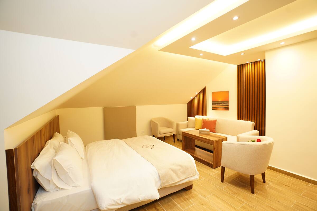 A modern hotel room with a sloped ceiling, a large bed with white linens, a seating area with a chair and a small table, and a wooden floor.