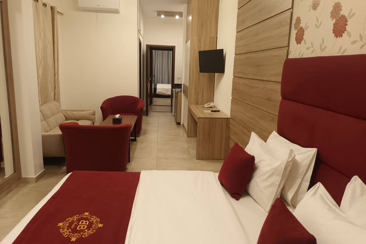 A hotel room with a large bed, red and white bedding, a wooden headboard, a small desk with a phone, a TV on the wall, and a sofa.