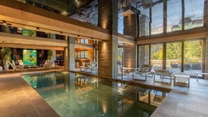 Zona de piscina interior àmplia amb parets de vidre transparent, bigues de fusta i mobiliari modern.