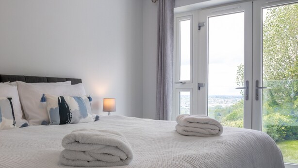 A neatly made bed with a white comforter and pillows, a stack of folded towels, and a view of greenery through the window. - Field View 5-bed House - Free Parking (Burton on Trent)