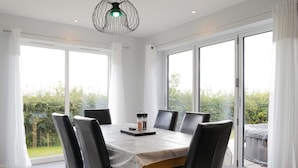 A dining area with a table set for six, surrounded by chairs, large windows, and a view of greenery. - Field View 5-bed House - Free Parking (Burton on Trent)