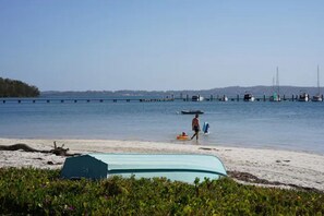A beach with a clear view of the water, a pier in the distance, and a boat docked near the shore. - Sunset Beach House (Soldiers Point)