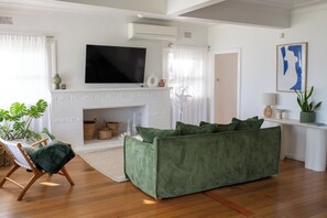 A living room with a green sofa, a flat-screen TV mounted above a fireplace, and a large window with sheer curtains. - Sunset Beach House (Soldiers Point)
