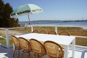 A deck with a white table and wicker chairs, overlooking a beach and pier. - Sunset Beach House (Soldiers Point)