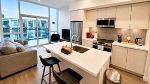 A modern kitchen with a large island, stainless steel appliances, and a view of a cityscape through large windows. - Stay in the heart of Friday Harbour! Cozy 1BR + sofa bed, steps from the Marina, Promenade, dining, shops and resort activities. (Innisfil)