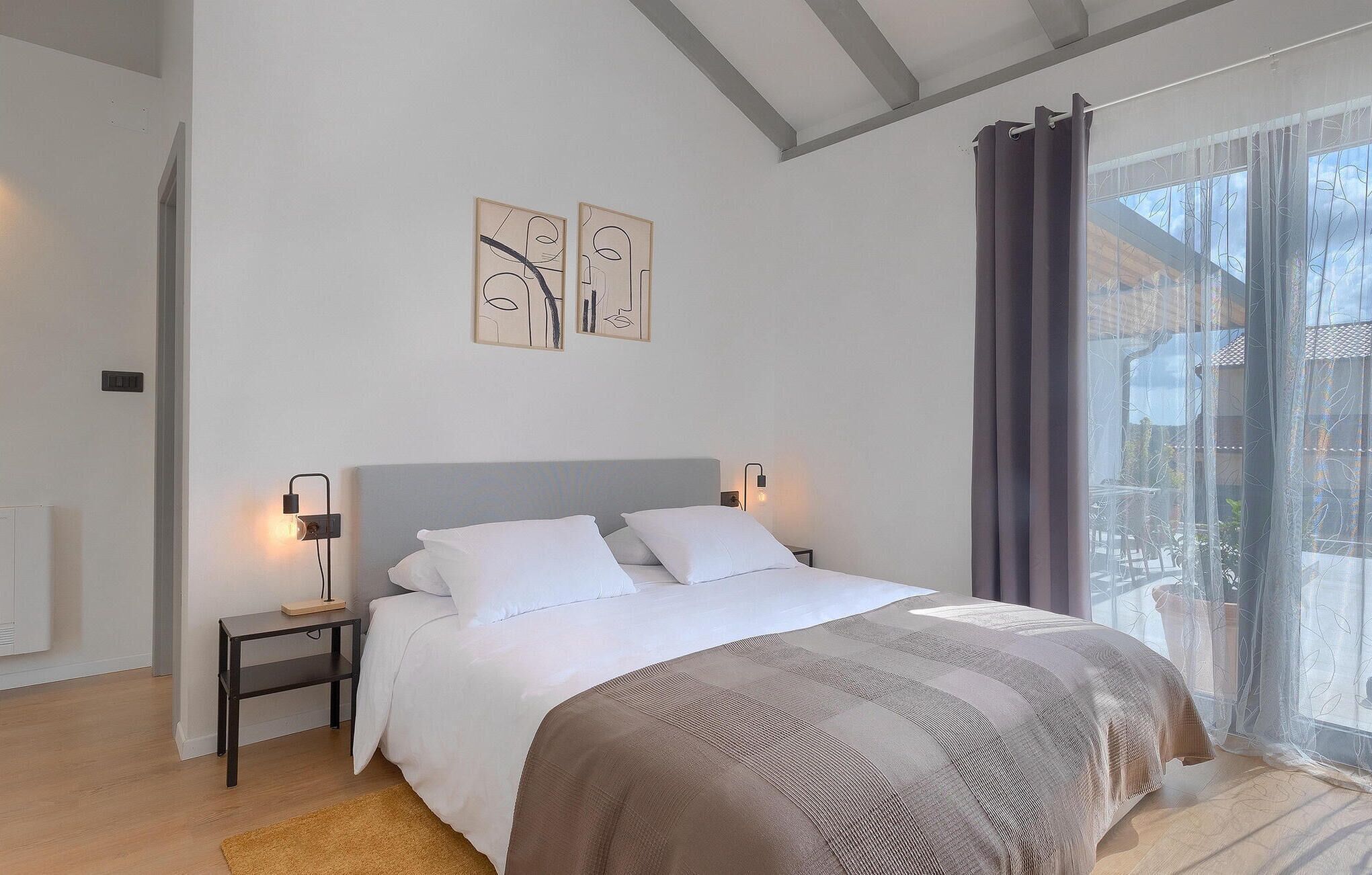 A bedroom with a bed, bedside tables, artwork, and a view through a window.