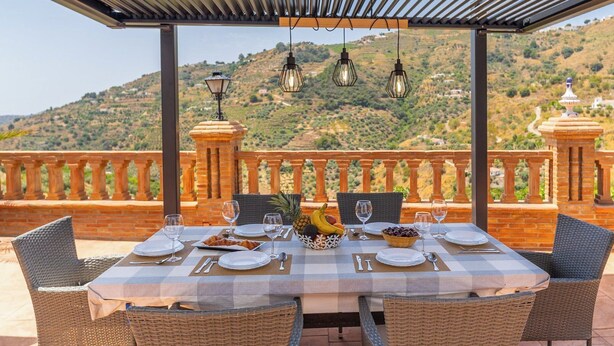 An outdoor dining area with a checkered tablecloth, wicker chairs, and a view of a hilly landscape. - Villa Almijara Cómpeta by Ruralidays (Málaga)