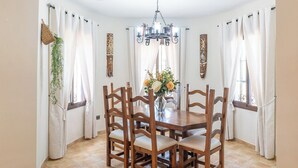 A dining area with a wooden table and chairs, white curtains, a chandelier, and a vase with flowers. - Villa Almijara Cómpeta by Ruralidays (Málaga)