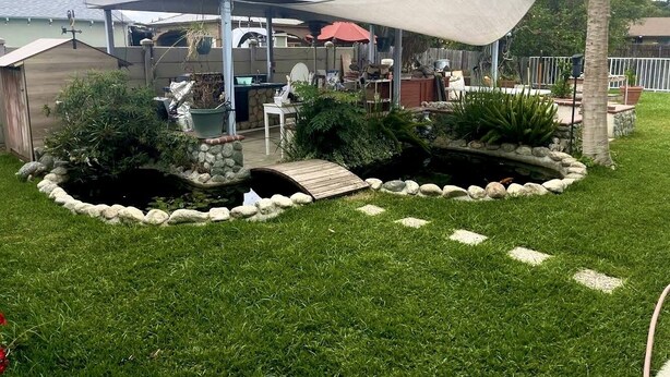 Ein Garten mit überdachter Terrasse, einem kleinen Teich mit Holzbrücke und einer Rasenfläche.