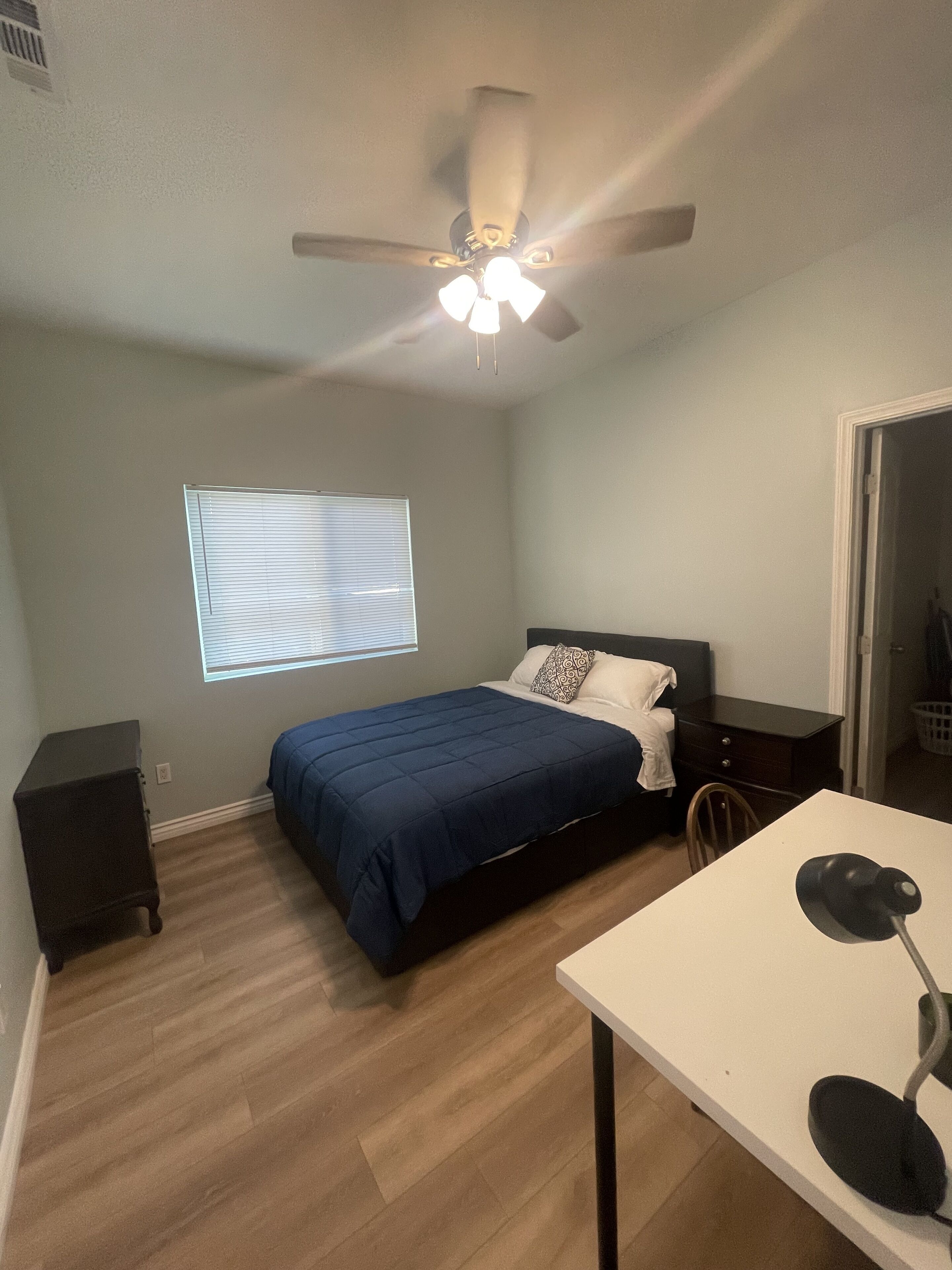 A bedroom with a bed, a desk, a chair, a ceiling fan, and a window with blinds.