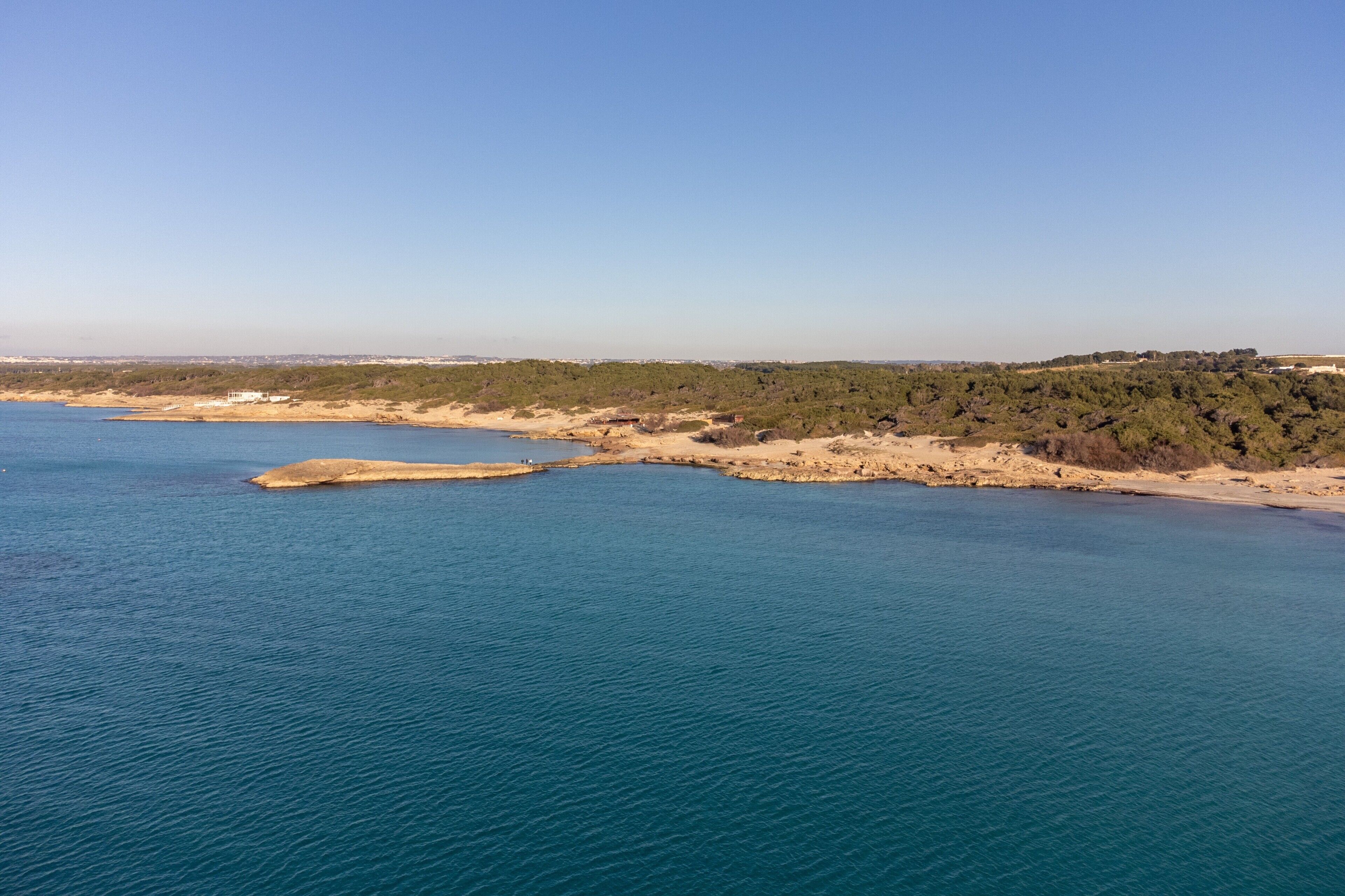 A coastal landscape with a sandy beach, clear blue sea, and dense greenery.