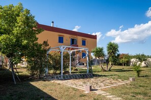 Edifici de dues plantes amb teulada vermella, zona exterior coberta i jardí amb arbres i un camí pavimentat.