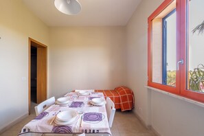 Un comedor con una mesa dispuesta para cuatro personas, una ventana con detalles rojos y una puerta que conduce a otra habitación.