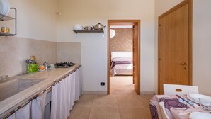 Eine schmale Küche mit Arbeitsplatte, Herd und Spüle. Eine Holztür führt zu einem Schlafzimmer mit Bett.