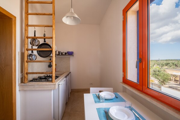 A kitchen with a gas stove, a wooden ladder shelf, hanging pots, a window with an orange frame, and a table set for two.